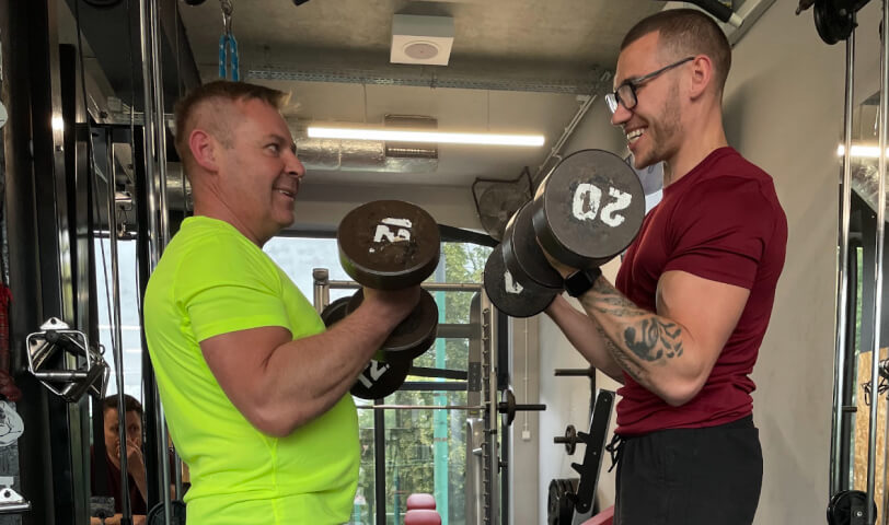 Trener personalny Robert Szałas i jego podopieczny uśmiechają się do siebie, jednocześnie wykonując uginanie ramion z hantlami na biceps.