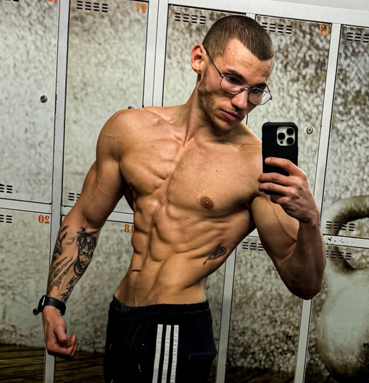 Robert Szałas, trener personalny, uśmiechnięty i umięśniony, pozuje przed lustrem w szatni, prezentując wyrzeźbioną sylwetkę.