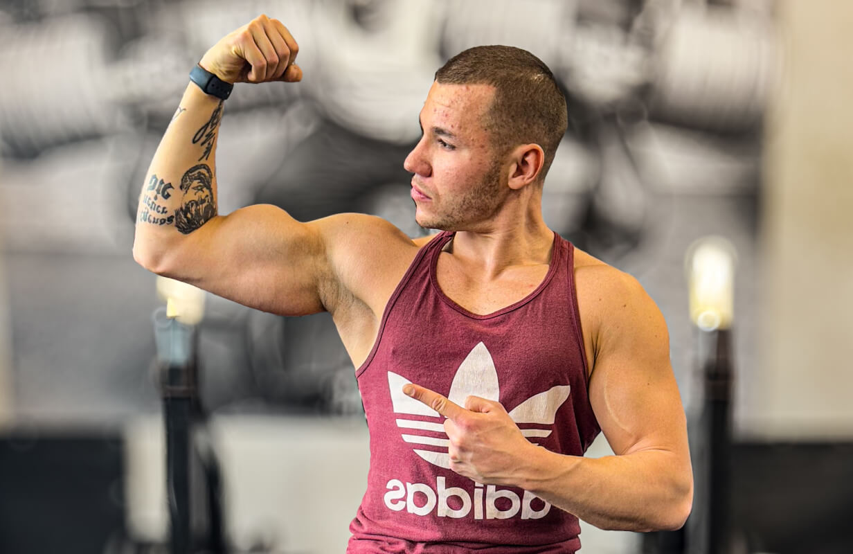 Trener personalny Robert Szałas napina biceps i wskazuje w lewo, zachęcając do przejścia na stronę z treningami personalnymi lub planami online.