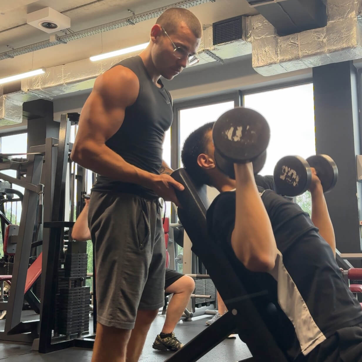 Trener personalny Robert Szałas asekuruje i nadzoruje technikę swojego podopiecznego podczas wyciskania hantli na barki w pozycji siedzącej.